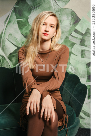 Young woman poses elegantly on green velvet couch with tropical backdrop in modern interior 125386696