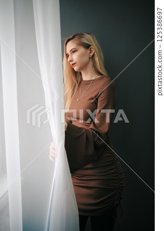 A young woman stands by a window with sheer curtains, looking out contemplatively A young woman stands by a window with sheer curtains, looking out contemplatively 125386697