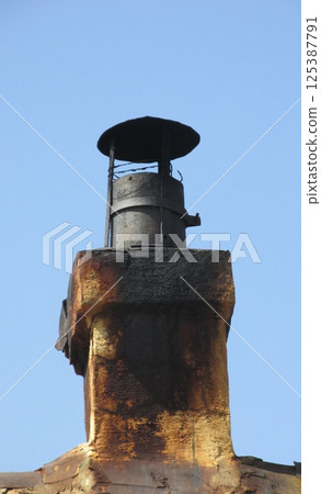 An old smoked chimney with a metal canopy against a blue sky An old smoked chimney with a metal canopy against a blue sky 125387791