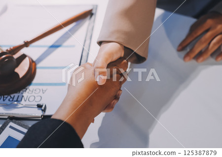 Two businesspeople are shaking hands in a law office with a scale and a gavel on the table 125387879