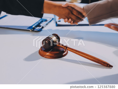 Two businesspeople are shaking hands in a law office with a scale and a gavel on the table 125387880