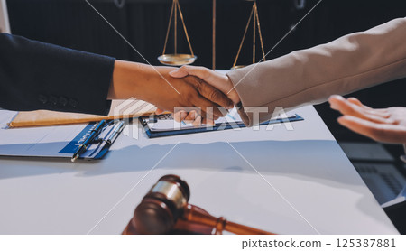 Two businesspeople are shaking hands in a law office with a scale and a gavel on the table 125387881