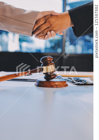 Two businesspeople are shaking hands in a law office with a scale and a gavel on the table 125387882