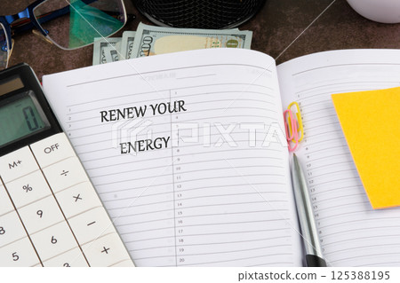 A planner open to a page featuring the phrase renew your energy is surrounded by a calculator, stationery, cash, and glasses, reflecting a productive workspace atmosphere 125388195
