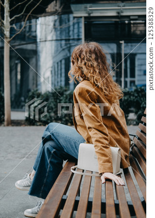 A young woman with curly red hair sits on a wooden bench smiling during a break in the city. Urban downtime, joyful pause, mindful living, mid-day rest... 125388329
