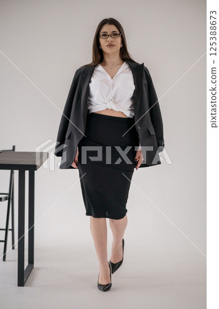 Professional woman with glasses walking in modern office space wearing black suit and knotted white shirt 125388673