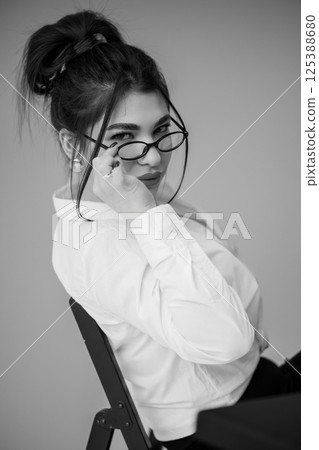 Portrait of a young woman with a serious expression, wearing glasses and a white shirt, seated on a chair Portrait of a young woman with a serious expression, wearing glasses and a white shirt, seated on a chair 125388680