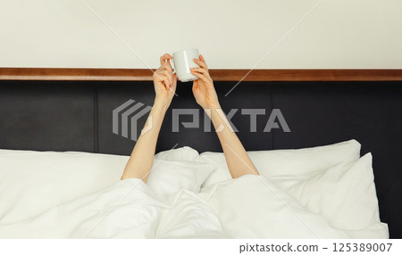 Cheerful lazy woman waking up after sleeping lying in bed holding cup in her hands asks for coffee 125389007