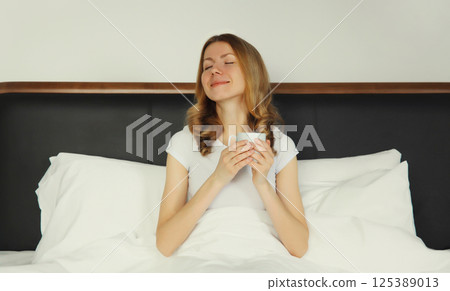 Happy smiling young woman enjoying with cup of hot coffee lying on bed at home 125389013
