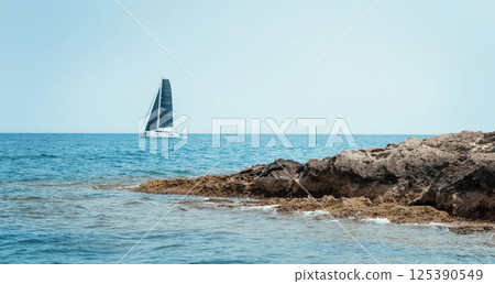 Seascape with sailboat behind island under blue cloudless sky. Nautical spirit and summer holidays Seascape with sailboat behind island under blue cloudless sky. Nautical spirit and summer holidays 125390549