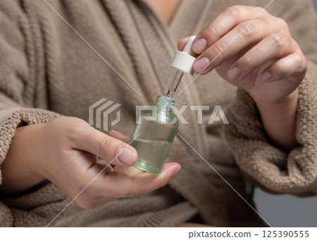 Closeup of woman in brown bathrobe opening green dropper bottle, cosmetic mockup Closeup of woman in brown bathrobe opening green dropper bottle, cosmetic mockup 125390555