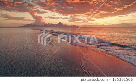 Aerial view capturing waves gently crashing on the shore of Tahiti at sunset, reflecting vibrant orange and red hues amidst a dramatic cloudscape 125391506