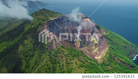 White smoke billowing from the crater of Mount Iliwerung volcano on Flores Island, Indonesia, surrounded by lush green vegetation and the blue ocean 125391615
