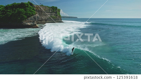 Aerial view of a surfer riding a wave near a cliff in Bali, Indonesia at Pantai Dreamland Beach, with other surfers waiting for their turn in the background, on a sunny day Aerial view of a surfer riding a wave near a cliff in Bali, Indonesia at Pantai Dreamland Beach, with other surfers waiting for their turn in the background, on a sunny day 125391618