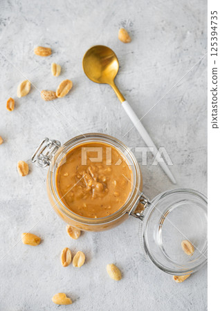 Glass jar with creamy peanut butter with spoon on grey background with scattered roasted nuts Glass jar with creamy peanut butter with spoon on grey background with scattered roasted nuts 125394735