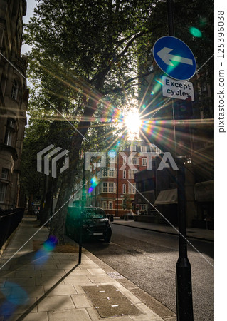 Sunlight shines through trees along a city street, with a road sign indicating a left turn for cycles. The urban scene features a quiet street with a parked vehicle and historic buildings in the 125396038