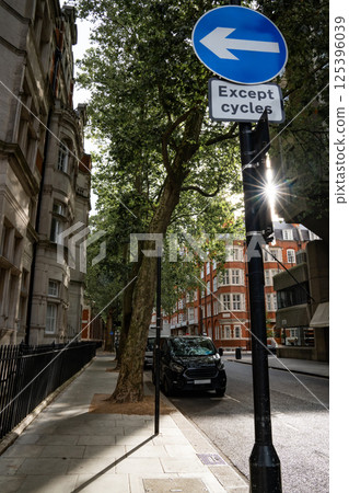 Sunlight shines through trees along a city street, with a road sign indicating a left turn for cycles. The urban scene features a quiet street with a parked vehicle and historic buildings in the 125396039