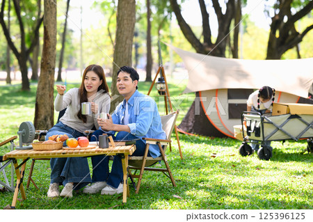 Asian couple enjoying hot drinks at a campsite while child plays in the background Asian couple enjoying hot drinks at a campsite while child plays in the background 125396125