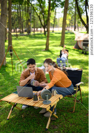 Happy couple enjoying hot drinks at a campsite while child plays in the background 125396130