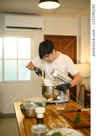 Young asian man in white shirt uses a stand mixer to whip cream in a cozy kitchen 125396298