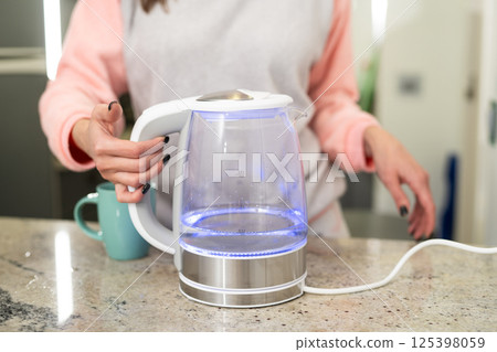 Woman in kitchen holding boiling modern glass electric kettle. Woman in kitchen holding boiling modern glass electric kettle. 125398059
