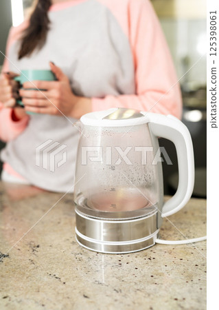 Modern glass electric kettle with boiling water. In the background there is a girl with a cup. Modern glass electric kettle with boiling water. In the background there is a girl with a cup. 125398061