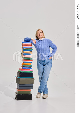Senior woman standing confidently next to high stack of colorful books, symbolizing academic success and years of accumulated knowledge. Senior woman standing confidently next to high stack of colorful books, symbolizing academic success and years of accumulated knowledge. 125399360