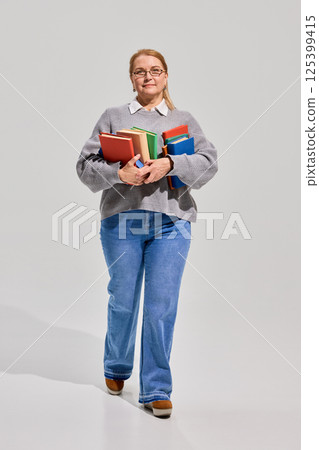 Portrait of elderly woman in casual clothes walking with stack of colorful books against white studio background. 125399415