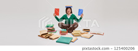 Happy elderly woman sitting on floor cross-legged, with book on head and two in hands, smiling against white studio background. 125400206