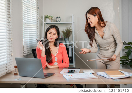 Two women working together in a bright office, discussing ideas with laptops and documents on the desk. 125401710