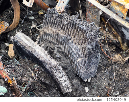 Burned heater core from a car showing severe fire damage, melted fins, and exposed internal structure lying among charred debris and wiring remnants 125402049