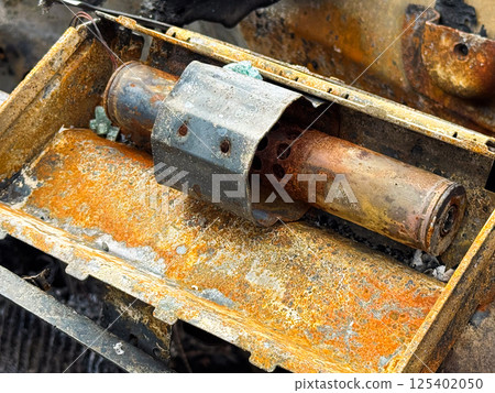 Burned steering column lock from a car with heavy rust and fire damage, showing the metal locking mechanism and oxidized surface inside a charred dashboard frame Burned steering column lock from a car with heavy rust and fire damage, showing the metal locking mechanism and oxidized surface inside a charred dashboard frame 125402050