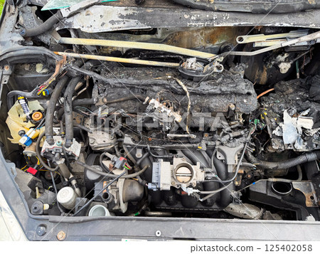 Burned car engine compartment showing fire-damaged engine block, melted wiring, scorched hoses, and exposed intake manifold with severe thermal destruction Burned car engine compartment showing fire-damaged engine block, melted wiring, scorched hoses, and exposed intake manifold with severe thermal destruction 125402058