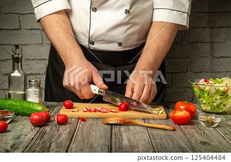 A chef is skillfully chopping vegetables on a wooden cutting board. Fresh tomatoes, radishes, and cucumber are present, showcasing vibrant colors in a cozy kitchen atmosphere 125404484