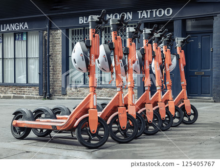 Bright orange electric scooters with white safety helmets parked on the sidewalk in downtown. 125405767