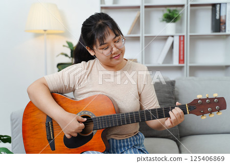 Happy Asian women play acoustic guitar and singing on the sofa in living room at home Happy Asian women play acoustic guitar and singing on the sofa in living room at home 125406869
