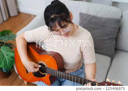 Happy Asian women play acoustic guitar and singing on the sofa in living room at home Happy Asian women play acoustic guitar and singing on the sofa in living room at home 125406873