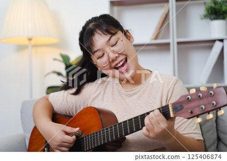 Happy Asian women play acoustic guitar and singing on the sofa in living room at home Happy Asian women play acoustic guitar and singing on the sofa in living room at home 125406874