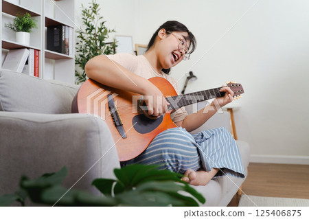 Happy Asian women play acoustic guitar and singing on the sofa in living room at home Happy Asian women play acoustic guitar and singing on the sofa in living room at home 125406875