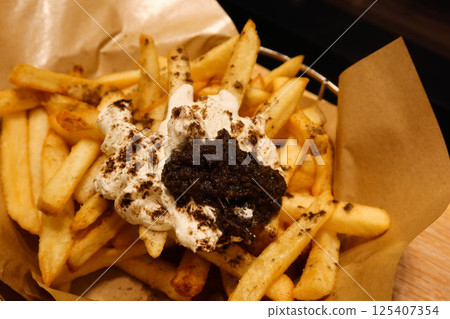 close up of a golden, crispy French fries presented in a rustic basket lined with brown paper. There is black truffle and whipped cream  as toppings. 125407354