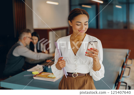 A Smiling Businesswoman Fully Engaged with Her Smartphone in a Sleek Modern Office Environment 125407640