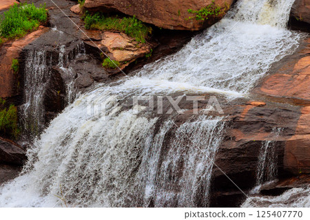 Rushing waterfall flows over smooth rocks in tranquil environment surrounded by greenery 125407770