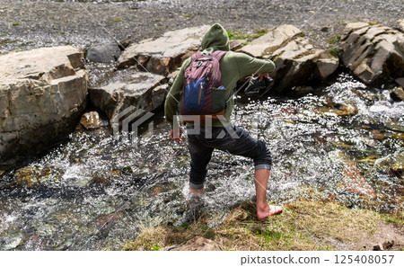 Young person wearing hoodie and backpack crosses rocky stream barefoot, holding shoes in hand Outdoor adventure and exploration in nature emphasizing self-reliance and balance, hiking and walking 125408057