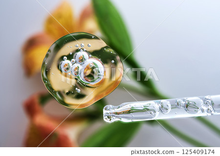 A pipette with a beautiful drop of serum on a background of lily flowers. A pipette with a beautiful drop of serum on a background of lily flowers. 125409174