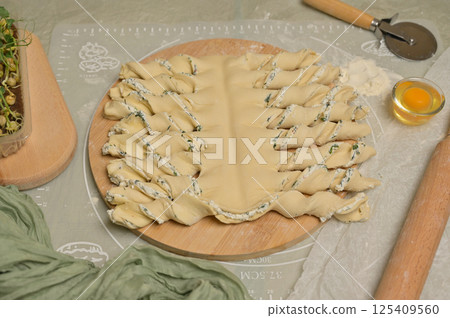 Twisted dough strips on wooden board. Cutter, egg, rolling pin, green towel, microgreens sprouts on kitchen table. Home cooking baking puffs 125409560