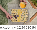 Top view woman's hands smears yolk on the twisted dough strips with a brush. Red small bowl with egg. Rolling pin, green towel, microgreens sprouts on kitchen table. Home cooking baking puffs 125409564