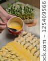 Close up woman's hands smears yolk on the twisted dough strips with a brush. Red small bowl with egg. Rolling pin, microgreens sprouts on kitchen table. Home cooking baking puffs 125409566