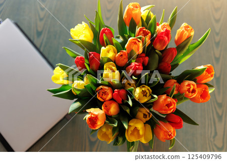 Top above view fresh multicolored tulips bouquet in vase table closed laptop warm sunset sun lights against balcony window cozy home interior. Blooming flowers decoration living room provence style Top above view fresh multicolored tulips bouquet in vase table closed laptop warm sunset sun lights against balcony window cozy home interior. Blooming flowers decoration living room provence style 125409796