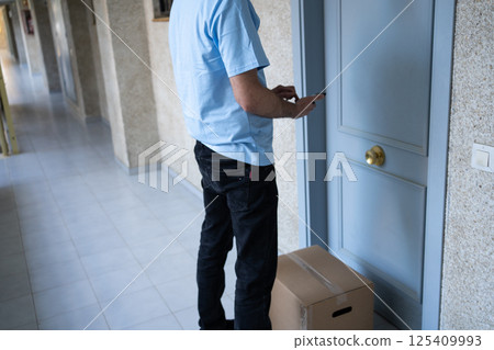 A male courier in a light blue uniform and cap delivers a package, standing in front of a residential apartment door. A male courier in a light blue uniform and cap delivers a package, standing in front of a residential apartment door. 125409993