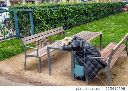 Vienna, Austria - 05.13.2023: Tired woman sleeps sitting at a table in one of the parks in Vienna. Vienna, Austria - 05.13.2023: Tired woman sleeps sitting at a table in one of the parks in Vienna. 125410396